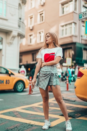 A woman stands confidently on an urban street, holding sunglasses in one hand. She is wearing a white T-shirt with a red design and a plaid skirt, paired with white sneakers. The background features yellow taxis, street signs, and tall buildings with various architectural styles.