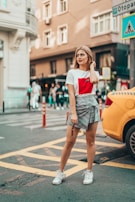 A stylish woman stands confidently on a city street. She wears a casual outfit consisting of a white t-shirt with a red design and a plaid skirt. People walk in the background while a yellow car is parked to her side. The urban environment features buildings and various street signs.