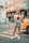 A stylish woman stands confidently on a city street. She wears a casual outfit consisting of a white t-shirt with a red design and a plaid skirt. People walk in the background while a yellow car is parked to her side. The urban environment features buildings and various street signs.