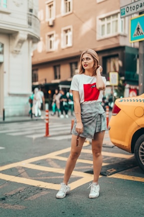A stylish woman stands confidently on a city street. She wears a casual outfit consisting of a white t-shirt with a red design and a plaid skirt. People walk in the background while a yellow car is parked to her side. The urban environment features buildings and various street signs.