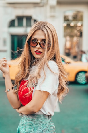 A stylish woman with long, wavy blonde hair and red lipstick is wearing sunglasses, a white t-shirt, a red handbag, and plaid pants. She stands confidently with one hand on her hip in an urban setting, with a blurry yellow car and buildings in the background.