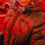 A person wearing a heavily padded, dark puffer jacket with a hood. The jacket has a zipper in the front and features a subtle embroidered logo on one sleeve. The person is also wearing a scarf and is standing outdoors, where a vehicle is partially visible in the background.