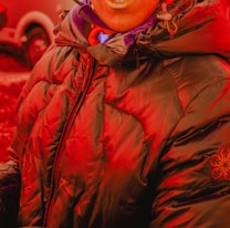 A person wearing a heavily padded, dark puffer jacket with a hood. The jacket has a zipper in the front and features a subtle embroidered logo on one sleeve. The person is also wearing a scarf and is standing outdoors, where a vehicle is partially visible in the background.