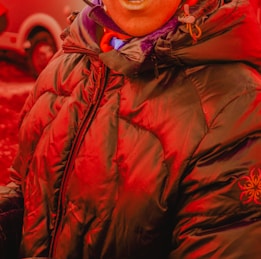 A person wearing a heavily padded, dark puffer jacket with a hood. The jacket has a zipper in the front and features a subtle embroidered logo on one sleeve. The person is also wearing a scarf and is standing outdoors, where a vehicle is partially visible in the background.
