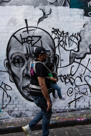 A man walks while carrying a child using a baby carrier in front of a large graffiti mural. The mural features a large, detailed face with abstract elements and tags surrounding it. The artwork covers a brick wall with various graffiti styles and forms.