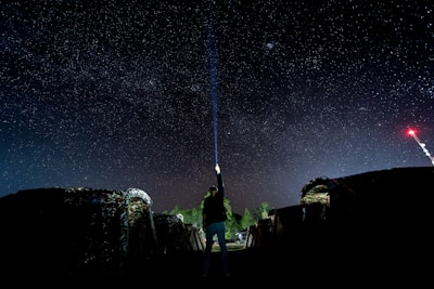 A person stands under a starry night sky holding a flashlight pointed upwards. The surroundings include tents and a radio tower with a red light in the distance, providing a sense of open nature and adventure.