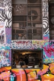A vibrant urban scene with a colorful graffiti-covered wall surrounds a small restaurant window where two chefs are seen preparing food inside. Industrial elements like grills and ventilation systems are visible above.