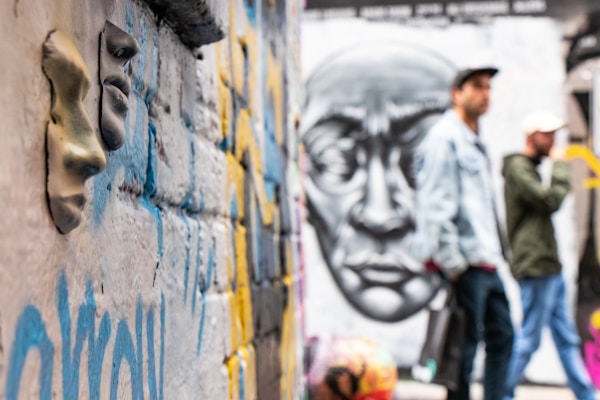 A textured brick wall features 3D face impressions alongside colorful graffiti. In the background, two indistinct figures stand with a large monochrome mural of a face on the wall behind them.