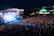 A vibrant outdoor concert scene features a large stage illuminated by bright lights, with a packed crowd of attendees gathered in front. A large overhead banner is visible, and a marquee tent is located to the right, also lit by colorful lights. The event appears to take place at night, with the skyline and distant lights visible in the background.