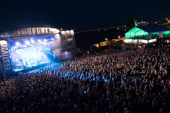 A vibrant outdoor concert scene features a large stage illuminated by bright lights, with a packed crowd of attendees gathered in front. A large overhead banner is visible, and a marquee tent is located to the right, also lit by colorful lights. The event appears to take place at night, with the skyline and distant lights visible in the background.