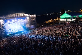 A vibrant outdoor concert scene features a large stage illuminated by bright lights, with a packed crowd of attendees gathered in front. A large overhead banner is visible, and a marquee tent is located to the right, also lit by colorful lights. The event appears to take place at night, with the skyline and distant lights visible in the background.