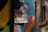 A piece of street art featuring a whimsical face made from recycled materials, including a tin can and bottle caps for eyes. Duct tape seems to hold the materials together against a vibrant graffiti-covered wall, with various colors and abstract patterns.