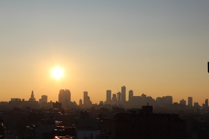 A cityscape silhouette under a rising or setting sun with a warm, glowing sky. The skyline features various high-rise buildings of different shapes against the gradient of orange and yellow hues stretching across the sky.