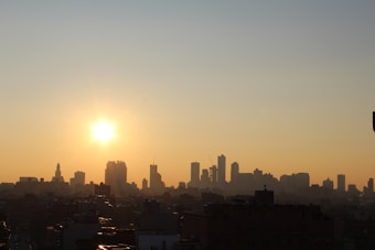 A cityscape silhouette under a rising or setting sun with a warm, glowing sky. The skyline features various high-rise buildings of different shapes against the gradient of orange and yellow hues stretching across the sky.