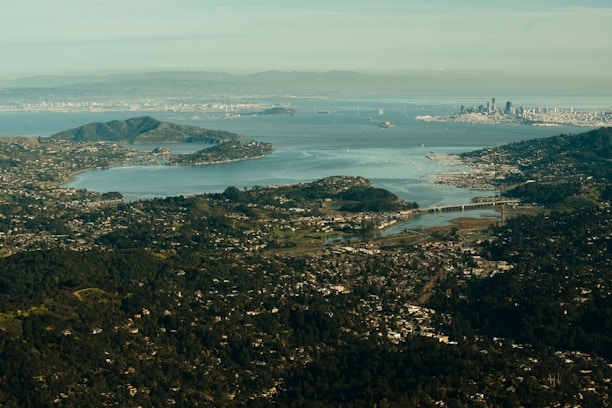 An aerial view of a bay area featuring a sprawling city skyline in the distance, numerous islands, and various waterways surrounded by greenery. The foreground shows a residential area with clusters of houses, while the backdrop displays distant mountains under a clear sky.