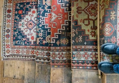 A series of intricately patterned carpets with geometric and floral designs in vibrant colors are laid on wooden steps. The patterns include interlocking shapes and stylized motifs. A pair of black shoes is partially visible standing on one of the steps, adding a human element to the scene.