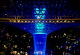 A striking indoor waterfall is illuminated with a vibrant blue light, cascading from a large, geodesic dome structure above, surrounded by lush greenery and architectural elements.