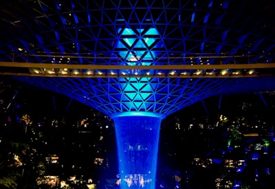 A striking indoor waterfall is illuminated with a vibrant blue light, cascading from a large, geodesic dome structure above, surrounded by lush greenery and architectural elements.