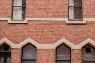 A facade of a building made of red brick with symmetrical architectural details. The structure includes two rectangular windows near the top and three pointed arch windows below. Cream-colored accents highlight the brickwork, and sheer curtains are visible in the upper windows.