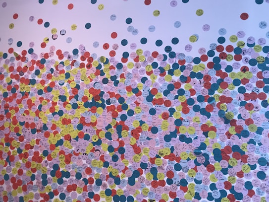 A wall densely covered with colorful circular stickers, each containing handwritten messages, drawings, and symbols. The stickers are predominantly in shades of red, yellow, blue, and white, and they create a lively and chaotic pattern. Various doodles, smiley faces, and short phrases are visible.