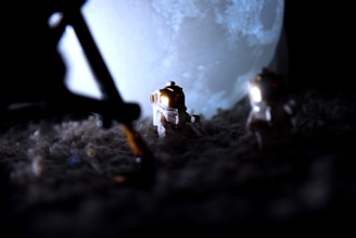 Two small astronaut figures stand on a textured surface resembling a lunar landscape, partially obscured by shadows. In the background, a large, illuminated sphere represents a planet or moon, casting a soft glow over the scene.