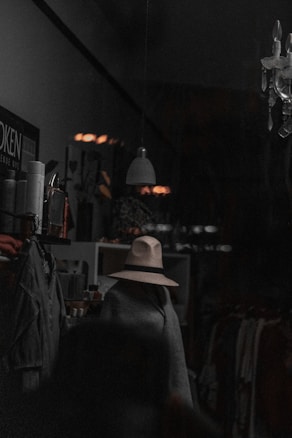 A dimly lit interior space with a mannequin wearing a beige hat and covered by a dark drape or cloak. There are various items on shelves, including clothes, spray bottles, and miscellaneous objects. A hanging light fixture is present in the foreground, and a chandelier light fixture is partially visible in the top right corner.