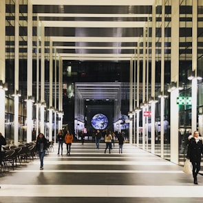 A modern indoor walkway lined with glass walls and bright overhead lighting. People are casually walking along, and a large, illuminated globe is positioned at the far end, creating a striking visual focal point.