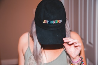 A person with long hair wearing a black cap with the word 'ASTROWORLD' on it in colorful letters. The person is holding the cap with one hand, tilting it slightly downward. They are dressed in a casual, sleeveless top and have several colorful bracelets on their wrist.