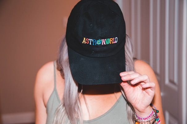 A person with long hair wearing a black cap with the word 'ASTROWORLD' on it in colorful letters. The person is holding the cap with one hand, tilting it slightly downward. They are dressed in a casual, sleeveless top and have several colorful bracelets on their wrist.