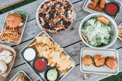 An array of various foods is spread out on a rustic wooden table. A pizza with charred toppings, a bowl of salad with cheese, a tray of nachos with dips including guacamole, salsa, and ranch, a hamburger with fries, sliders, and fried stick appetizers are neatly arranged. The scene conveys a casual dining setup, ideal for sharing.