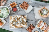 A variety of appetizing dishes are arranged on a rustic wooden table. There are trays of nachos topped with jalapeños and melted cheese, a bowl of fresh green salad with grated cheese, tacos filled with crispy ingredients, and a baked tortilla bowl with savory filling. Additional trays contain crispy fried items like mozzarella sticks and small fried dough balls, accompanied by dipping sauce.