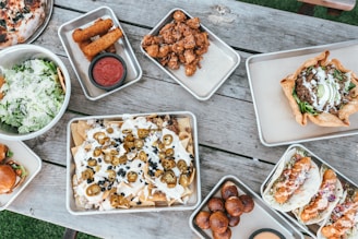 A variety of appetizing dishes are arranged on a rustic wooden table. There are trays of nachos topped with jalapeños and melted cheese, a bowl of fresh green salad with grated cheese, tacos filled with crispy ingredients, and a baked tortilla bowl with savory filling. Additional trays contain crispy fried items like mozzarella sticks and small fried dough balls, accompanied by dipping sauce.