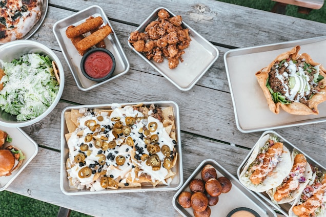 A variety of appetizing dishes are arranged on a rustic wooden table. There are trays of nachos topped with jalapeños and melted cheese, a bowl of fresh green salad with grated cheese, tacos filled with crispy ingredients, and a baked tortilla bowl with savory filling. Additional trays contain crispy fried items like mozzarella sticks and small fried dough balls, accompanied by dipping sauce.