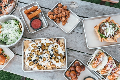 A variety of appetizing dishes are arranged on a rustic wooden table. There are trays of nachos topped with jalapeños and melted cheese, a bowl of fresh green salad with grated cheese, tacos filled with crispy ingredients, and a baked tortilla bowl with savory filling. Additional trays contain crispy fried items like mozzarella sticks and small fried dough balls, accompanied by dipping sauce.