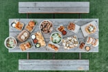 A variety of food items are arranged on metal trays atop a rustic wooden table. The spread includes a pizza with olives, two burgers with fries, assorted dips, nachos with jalapenos and olives, tacos, fried appetizers, salad bowls, and a few rolls. This setup is on a green grass surface with wooden benches on either side.