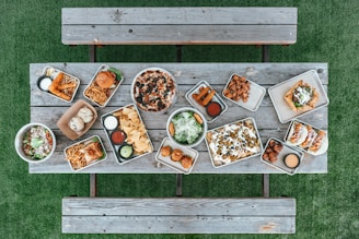 A variety of food items are arranged on metal trays atop a rustic wooden table. The spread includes a pizza with olives, two burgers with fries, assorted dips, nachos with jalapenos and olives, tacos, fried appetizers, salad bowls, and a few rolls. This setup is on a green grass surface with wooden benches on either side.