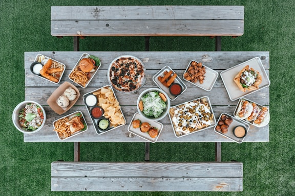 A variety of food items are arranged on metal trays atop a rustic wooden table. The spread includes a pizza with olives, two burgers with fries, assorted dips, nachos with jalapenos and olives, tacos, fried appetizers, salad bowls, and a few rolls. This setup is on a green grass surface with wooden benches on either side.