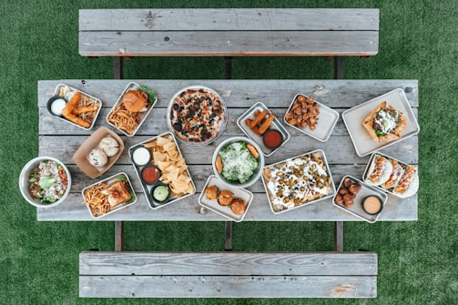 A variety of food items are arranged on metal trays atop a rustic wooden table. The spread includes a pizza with olives, two burgers with fries, assorted dips, nachos with jalapenos and olives, tacos, fried appetizers, salad bowls, and a few rolls. This setup is on a green grass surface with wooden benches on either side.