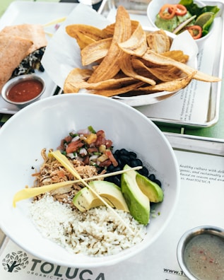 A bowl containing rice, shredded meat, avocado slices, black beans, pico de gallo, and crumbled cheese is presented on a tray. Nearby, a bowl of plantain chips is accompanied by small dishes of sauce. A wrap filled with greens and vegetables is placed alongside, with a side of salsa. Also included on the tray is a bowl of salad with sliced cherry tomatoes.