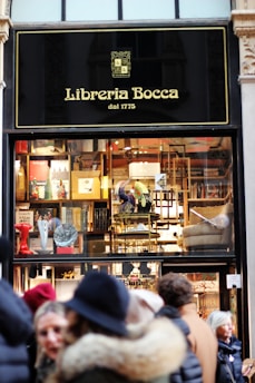 A storefront of a bookstore named Libreria Bocca, established in 1775, featuring a window display with various books and decorative items. The exterior sign is black with gold lettering. Several people in winter attire are standing outside.