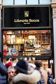 A storefront of a bookstore named Libreria Bocca, established in 1775, featuring a window display with various books and decorative items. The exterior sign is black with gold lettering. Several people in winter attire are standing outside.