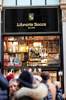 A storefront of a bookstore named Libreria Bocca, established in 1775, featuring a window display with various books and decorative items. The exterior sign is black with gold lettering. Several people in winter attire are standing outside.