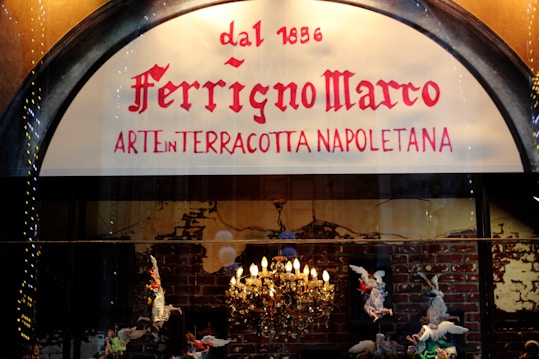 A shop sign displays the name 'Ferrigno Marco' with the phrase 'Arte in Terracotta Napoletana' below. The background features a lit chandelier and terracotta figurines of angels against a brick wall.