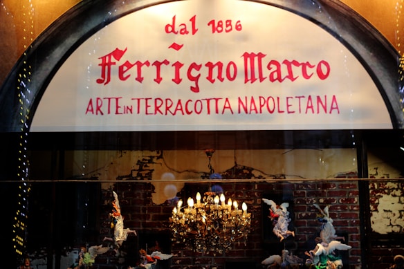 A shop sign displays the name 'Ferrigno Marco' with the phrase 'Arte in Terracotta Napoletana' below. The background features a lit chandelier and terracotta figurines of angels against a brick wall.