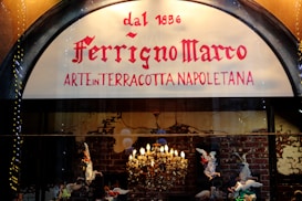 A shop sign displays the name 'Ferrigno Marco' with the phrase 'Arte in Terracotta Napoletana' below. The background features a lit chandelier and terracotta figurines of angels against a brick wall.