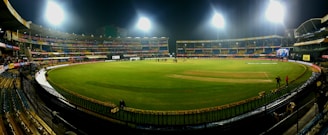 A brightly lit cricket stadium under the night sky with a lush green field, surrounded by multicolored seating and empty stands. A few people can be seen on the field, possibly players or staff.