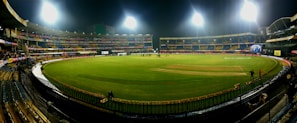 A brightly lit cricket stadium under the night sky with a lush green field, surrounded by multicolored seating and empty stands. A few people can be seen on the field, possibly players or staff.