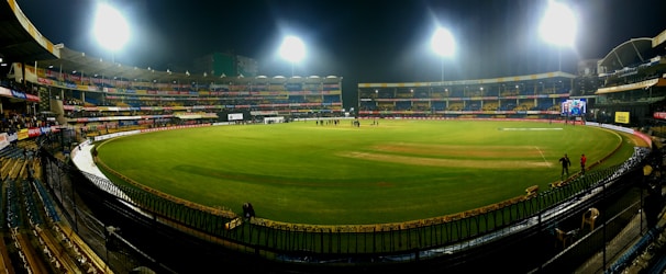 A brightly lit cricket stadium under the night sky with a lush green field, surrounded by multicolored seating and empty stands. A few people can be seen on the field, possibly players or staff.