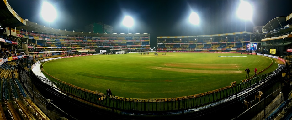 A brightly lit cricket stadium under the night sky with a lush green field, surrounded by multicolored seating and empty stands. A few people can be seen on the field, possibly players or staff.