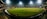 A brightly lit cricket stadium under the night sky with a lush green field, surrounded by multicolored seating and empty stands. A few people can be seen on the field, possibly players or staff.